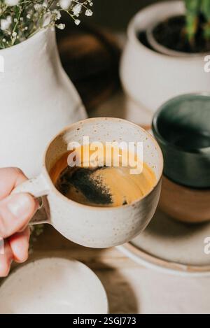 A person is holding a cup of coffee with a spoon in it. The coffee is brown and has a strong aroma. The scene is set on a table with a vase and a pott Stock Photo