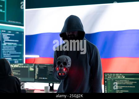 Conceptual image of a hooded russian hacker creating hazardous alerts, holds a skull and warning of death threats. Russian federation works on cybercrime and violence, panic and torment. Stock Photo