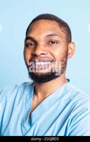 Portrait of african american male nurse smiling in front of camera, posing with confidence against blue background. Cheerful guy wearing protective clinical workwear to fight disease. Stock Photo