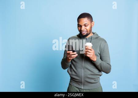 Confident optimistic man browsing his smartphone and serving coffee, feeling happy after receiving good news and socializing with his friends via text messages. Modern guy with a refreshment. Stock Photo