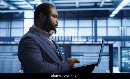 African american man walking in high tech facility, using laptop to patch network issues leading to slow data transfer rates. Data center worker fixing errors affecting supercomputers, camera A Stock Photo