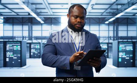 Software developer managing data center with advanced technology, ensuring efficient network management. Admin walking in server room, optimizing data center infrastructure with tablet, camera B Stock Photo