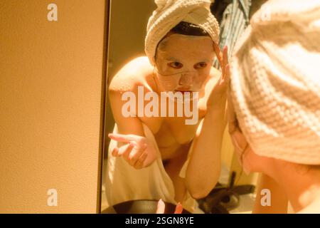 Woman applying a facial mask in front of a mirror with a towel wrapped around her head in a hotel, Tokyo, Japan Stock Photo