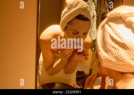 Woman in towel applies a face mask while looking in the mirror in a softly lit hotel bathroom. Tokyo, Japan Stock Photo
