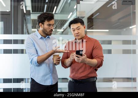Two colleagues debating and analyzing data on a smartphone while standing in a professional workspace Stock Photo