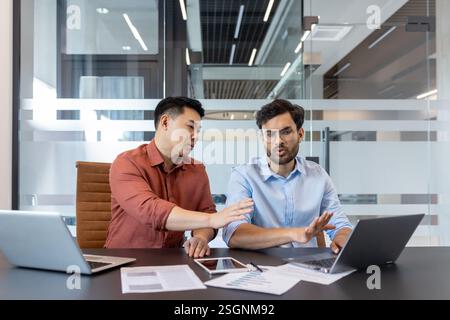 Two colleagues in casual clothes discussing a work project, men sitting at a desk with a laptop. Viewing electronic online financial reports. Stock Photo