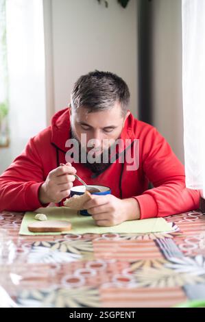 Indoor shot of a bearded man in a red jacket, focusing intently on his soup, with bread and a patterned table adding warmth. Natural light filters in, Stock Photo