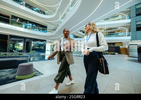 Two professional women are walking through a contemporary office environment, engaging in a lively conversation. Teamwork, communication, and modern c Stock Photo