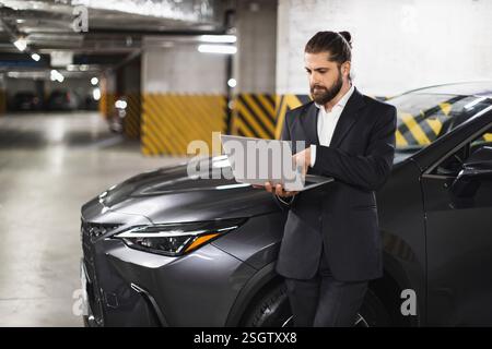 Caucasian male businessman using laptop in parking garage beside modern vehicle. Professional setting, focused on work, wearing stylish suit Stock Photo
