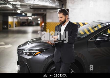 Caucasian male businessman using laptop in parking garage beside modern vehicle. Professional setting, focused on work, wearing stylish suit Stock Photo
