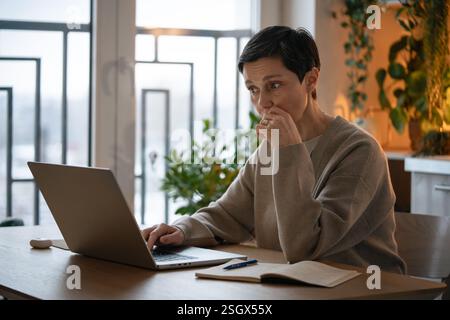 Meeting deadlines. Thoughtful worried mature woman working on laptop, taking notes at home alone Stock Photo