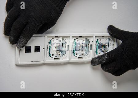 Close-up of an electrician wearing black gloves installing power socket on white wall. Electrical repair and maintenance process in progress Stock Photo