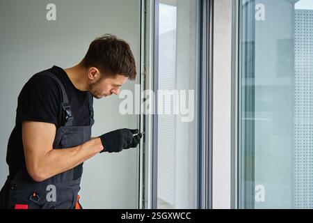 Skilled worker wearing gloves uses screwdriver to fix window frame, home renovation tasks. Professional repairman adjusting white pvc windows in moder Stock Photo