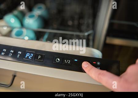 Close-up of female finger pressing button on dishwasher control panel. Woman using dishwasher in modern kitchen. Modern kitchen appliance with digital Stock Photo