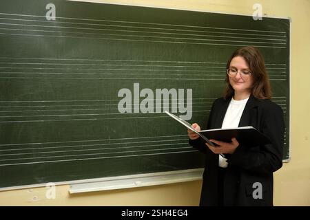 A smiling woman, who is a teacher, stands in front of the blackboard, engaging students in learning. The happy teacher is standing by the chalkboard, Stock Photo