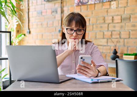 Portrait off middle-aged business woman working with laptop, using phone, sitting at desk Stock Photo