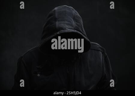 Unrecognizable cyber criminal or computer hacker wearing black hooded jacket, low key portrait, selective focus Stock Photo