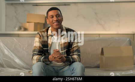 Joyful ethnic African American man guy tenant sit on couch sofa smiling happy male homeowner enjoy moving in new home relocation buying apartment rent Stock Photo