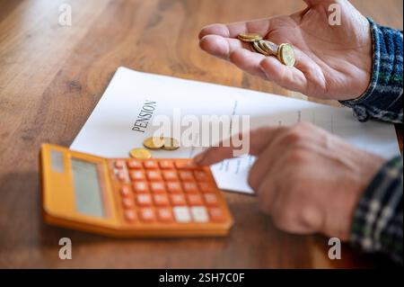 An elderly person counts coins while using a calculator, reviewing a pension document on a wooden table, symbolizing financial planning and retirement Stock Photo