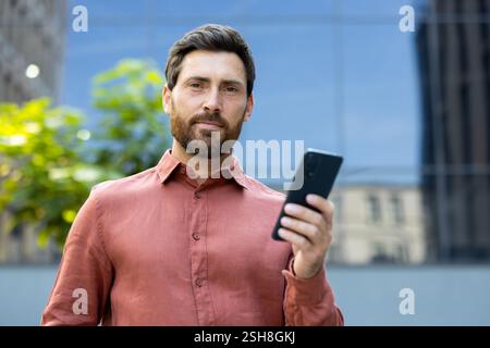 A confident man casually holding a modern smartphone, set prominently against a bustling and vibrant city backdrop. Stock Photo