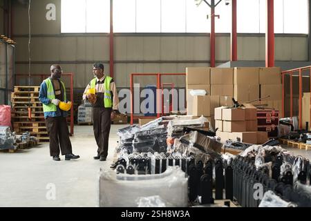 Two intercultural male workers of warehouse or factory discussing working points while moving along aisle between stacks of items Stock Photo