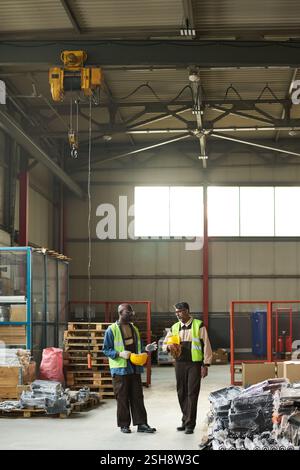 Two male workers of modern factory discussing quality of equipment while standing on aisle between stacks of packed items Stock Photo