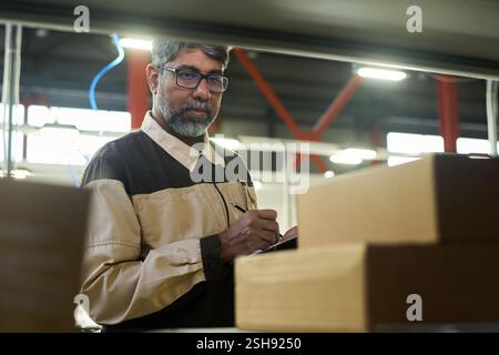 Mature multiethnic factory worker taking notes while looking at sticker on packed cardboard box during revision in warehouse Stock Photo