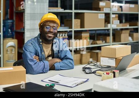 Happy young African American male engineer in hardhat and workwear looking at camera while sitting by his workplace Stock Photo