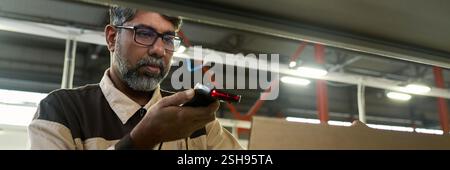 Header of mature multiethnic factory worker scanning barcodes on packed cardboard boxes while making revision in warehouse Stock Photo