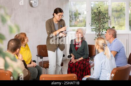 Young woman presides over support group meeting with mature people listening Stock Photo