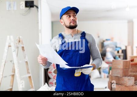 Responsible focused bearded supervisor wearing blue overall taking notes on papers while inspecting construction site Stock Photo
