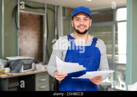 Responsible focused bearded supervisor wearing blue overall taking notes on papers while inspecting construction site Stock Photo