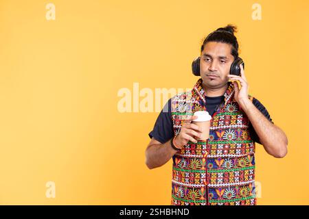 Tired indian man listening music, enjoying hot beverage. Person wearing headphones, hearing songs, drinking coffee to wake up and gain energy, isolated over studio background Stock Photo