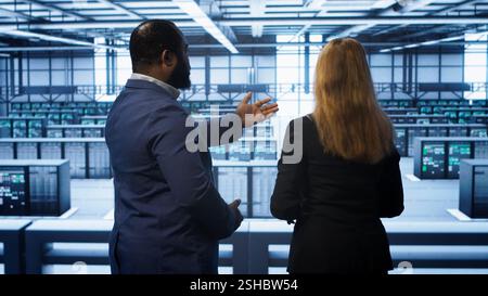 Data center engineer presenting equipment to inspector supervising operations. IT specialist in server farm doing tour for supervisor monitoring equipment performance, camera A Stock Photo