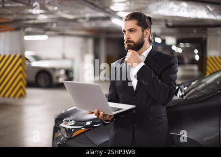 Caucasian male businessman in suit standing in parking garage using laptop, appearing thoughtful and focused. Middle-aged professional contemplating work decisions in modern business environment. Stock Photo