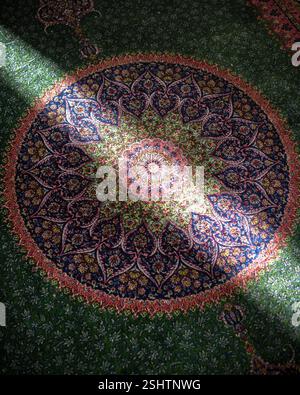 A circular Persian rug featuring an elaborate central medallion and radiating floral patterns illuminated by a strong beam of light Stock Photo
