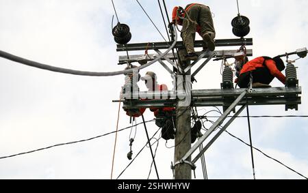 Two electrical workers in bright orange gear performing maintenance on power lines and utility poles under a cloudy sky Stock Photo