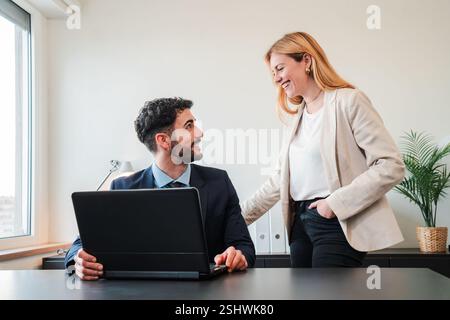 Cheerful Interaction Between Business Partners Working Collaboratively at a Laptop in a Bright Office. Engaged Man and Woman Share Ideas and Laugh Together, Creating a Positive and Productive Work. High quality photo Stock Photo