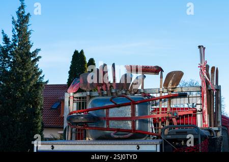 A variety of garden tools, including shovels and a wheelbarrow, are stacked on a trailer under a clear blue sky. This outdoor setup showcases readines Stock Photo