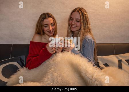 Two friends are relaxing on a sofa with a warm blanket, enjoying a coffee and browsing social media on a smartphone Stock Photo