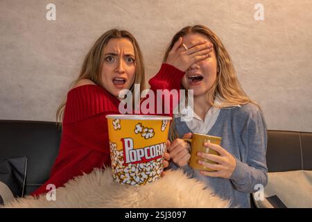 Two friends are watching a horror movie on television, eating popcorn and drinking from mugs Stock Photo