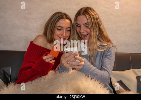 Two joyful female friends making online purchases using a smartphone and credit card, enjoying their shopping time together on a comfortable couch Stock Photo