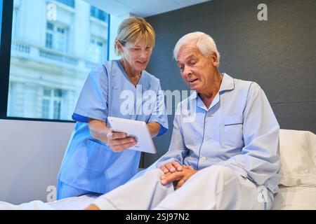 A senior man in hospital attire engages with a nurse as they review information on a digital tablet, providing insights in a healthcare setting. Stock Photo