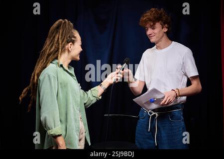 Smiling young man and woman sharing microphone on stage while looking at each other holding paper and wearing casual attire Stock Photo