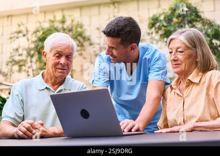 A healthcare worker helps two senior individuals navigate using a laptop in a pleasant outdoor setting, showcasing care, education, and technology sup Stock Photo
