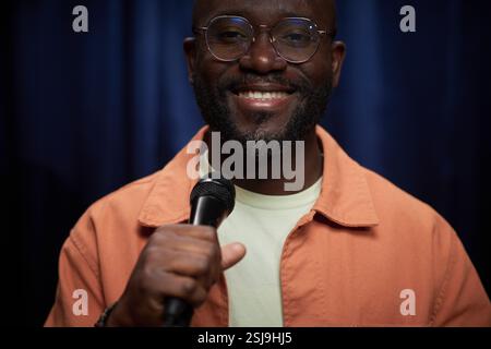 Portrait of African American man smiling while holding microphone on stage against dark background wearing glasses and orange jacket Stock Photo