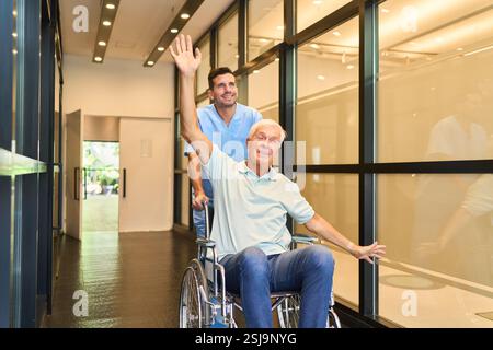 A cheerful senior man being assisted by a healthcare worker in a rehabilitation center hallway. The man in the wheelchair appears happy and is gesturi Stock Photo