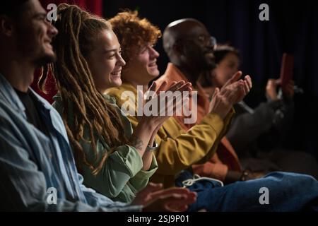 Group of diverse people sitting in row and applauding during a performance, capturing joy and engagement, showcasing various ethnic backgrounds and unity Stock Photo