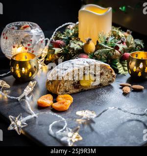 Christmas baking-Stollen loaf cut open showing marzipan on black tiled board with starred lights, decorations, candle, fruit, nuts in a square format Stock Photo