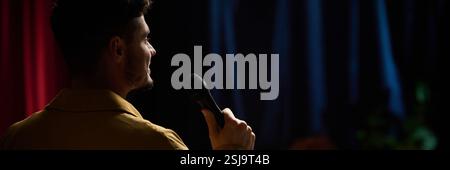 Comedian performing stand-up comedy on stage while holding microphone in hand, background curtains creating dramatic atmosphere Stock Photo